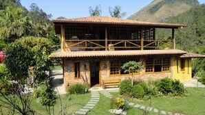Una casa de dos pisos con balcón, rodeada de exuberante vegetación y montañas al fondo.