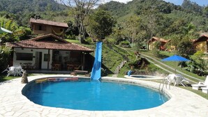 Una piscina con tobogán, rodeada por un patio de piedra, y un edificio con tejado de azulejos.