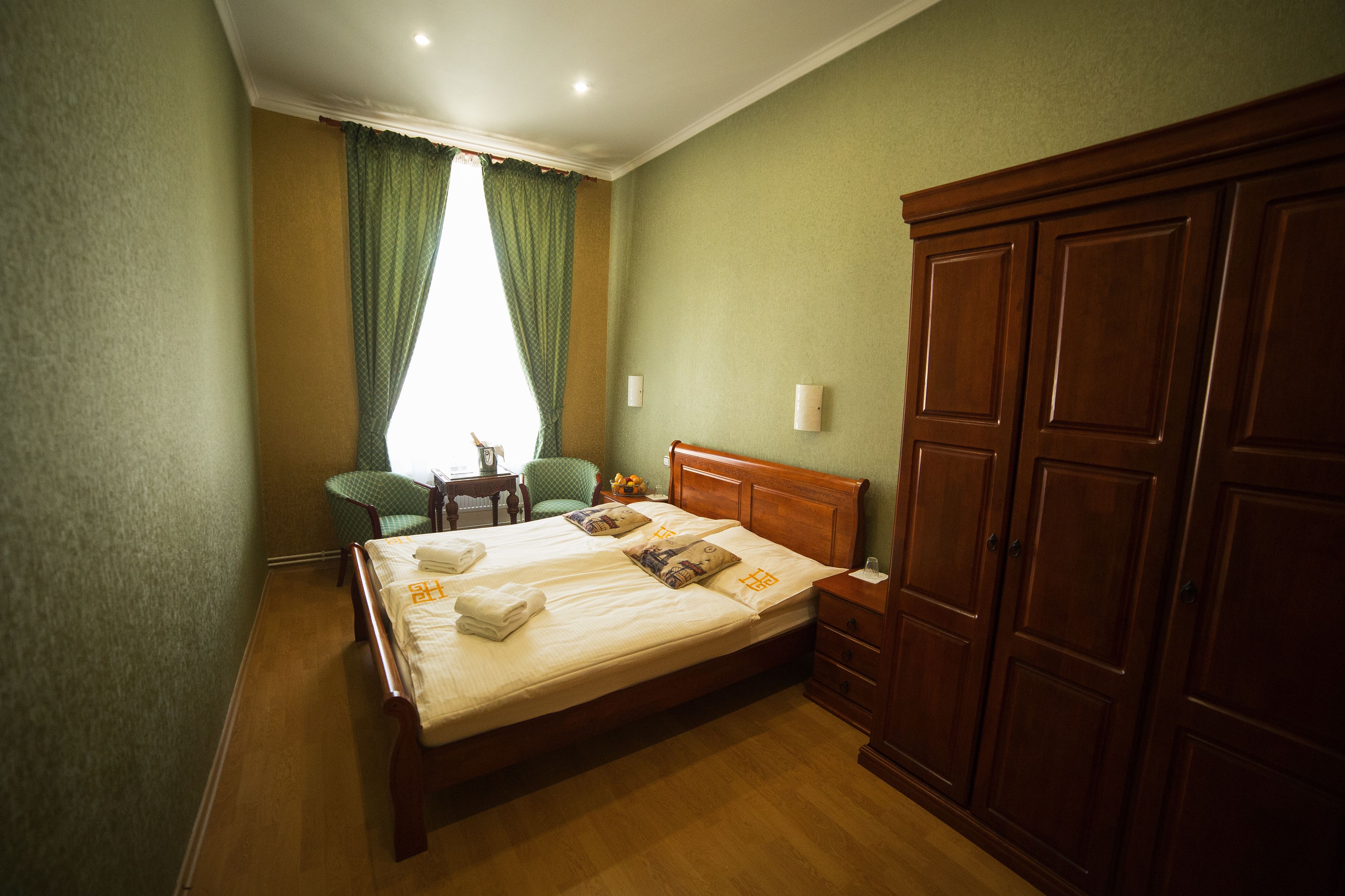 Ein ordentlich bezogenes Bett mit Kissen und Decke, ein Holzschrank, ein kleiner Tisch mit Stuhl und ein Fenster mit Vorhängen.