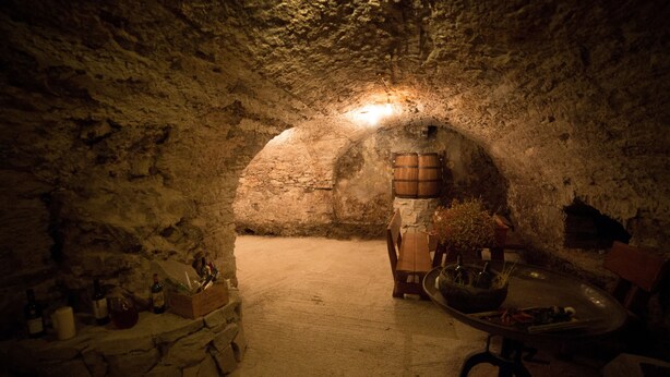 Una bodega subterránea tenuemente iluminada, con paredes de piedra, barriles de madera y una mesa rústica con un cuenco.