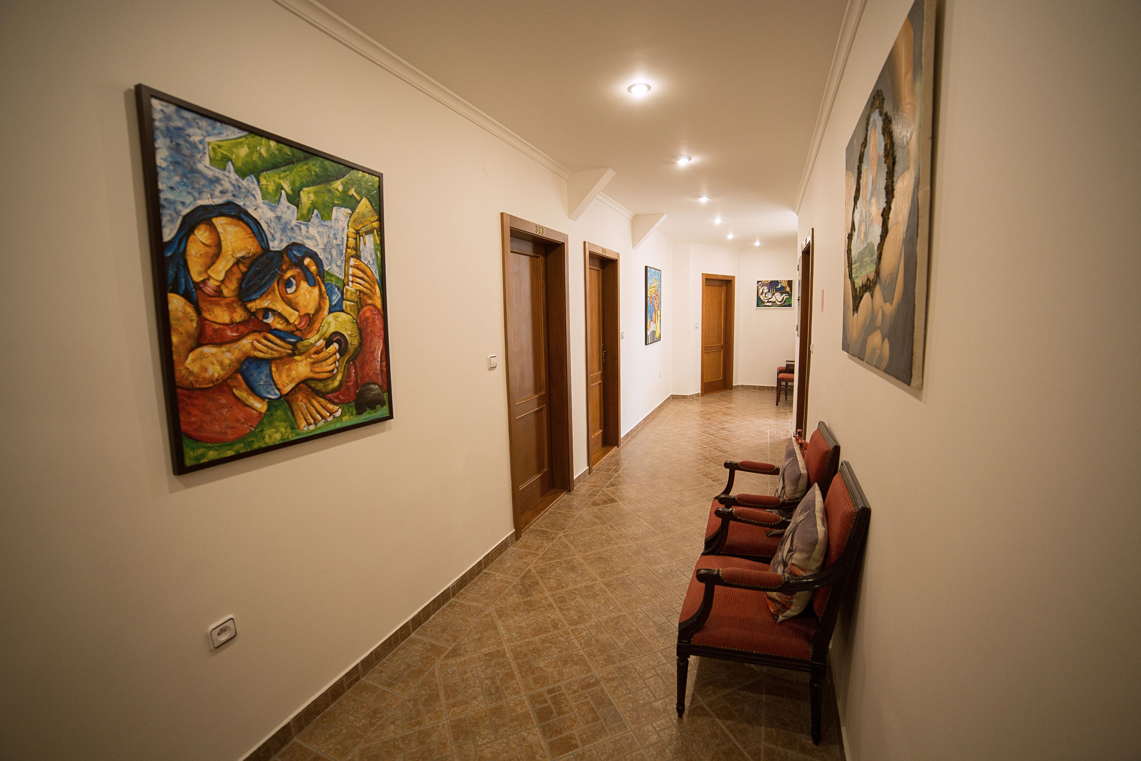 A hallway with a row of wooden doors, a bench with pillows, and a large colorful painting on the wall.