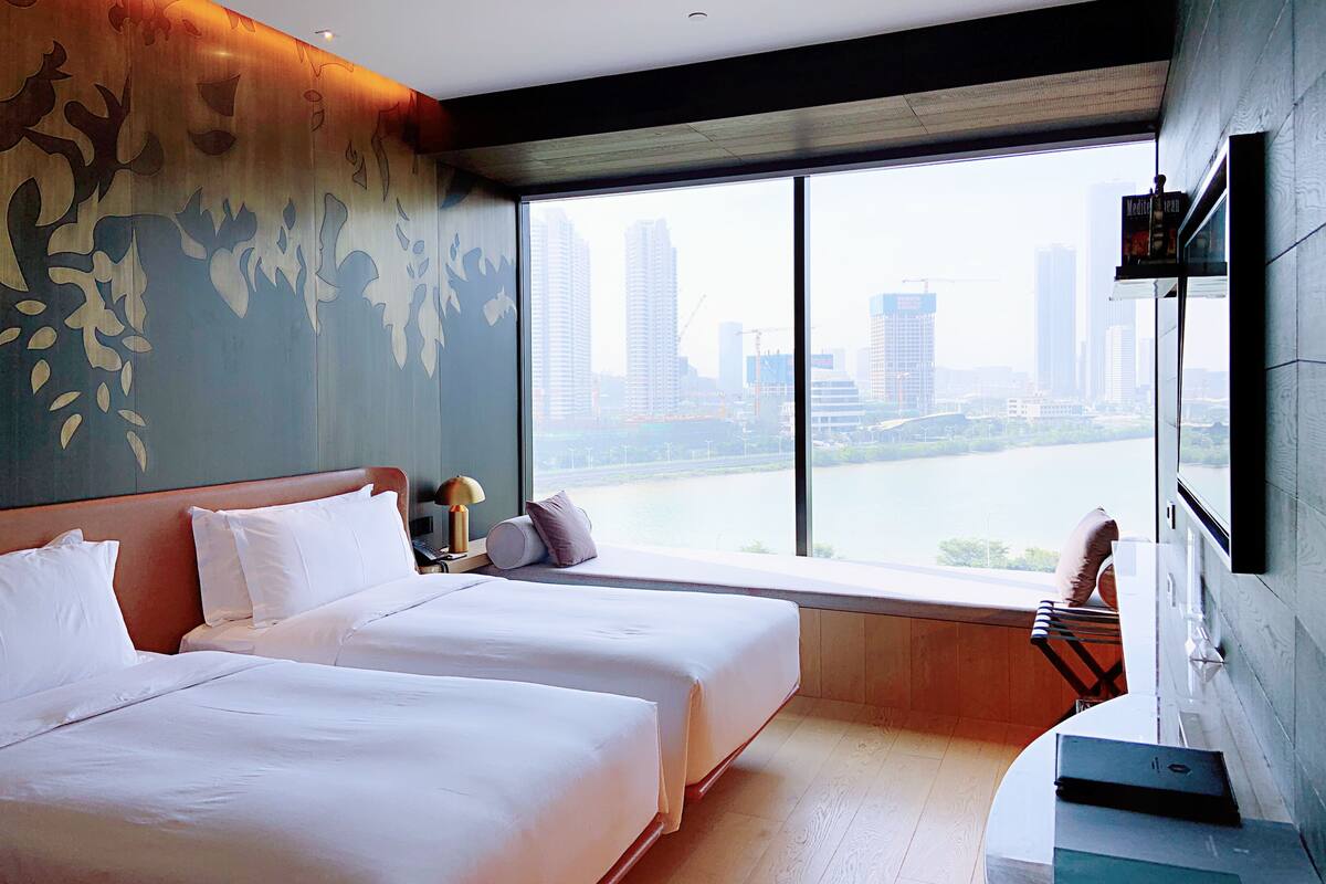 A hotel room with two beds, a large window with a city view, a TV, and a desk with a lamp.