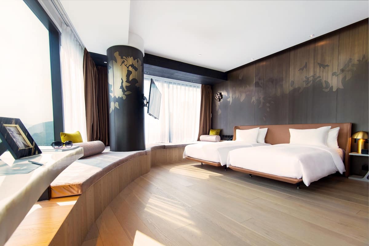 A modern hotel room with a curved desk, two beds, a large window, and a view of the city.