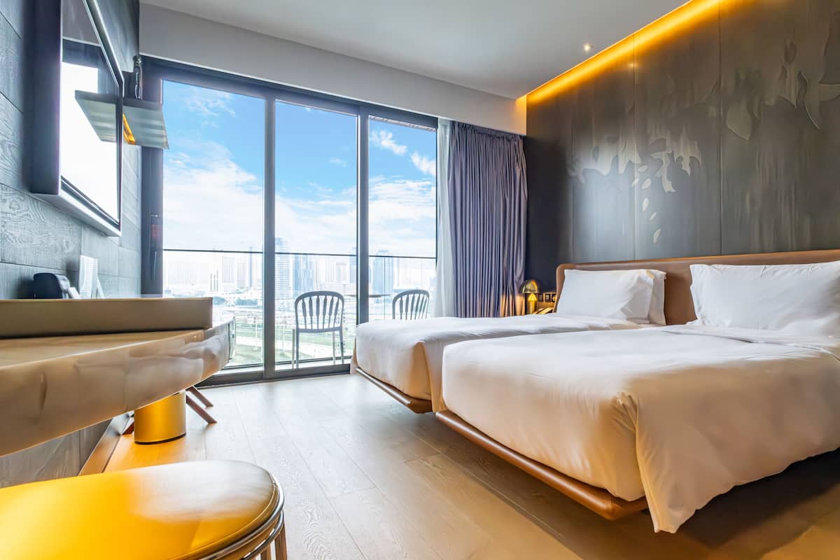 A modern hotel room with a large bed, a desk, and a view of the city skyline through floor-to-ceiling windows.