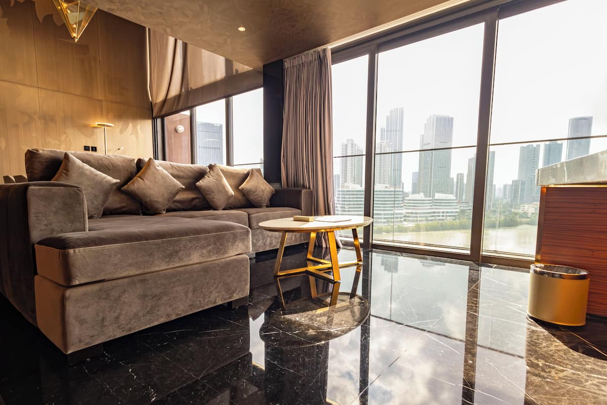 A modern living room with a grey sofa, a small round table, and a view of a cityscape through large windows.