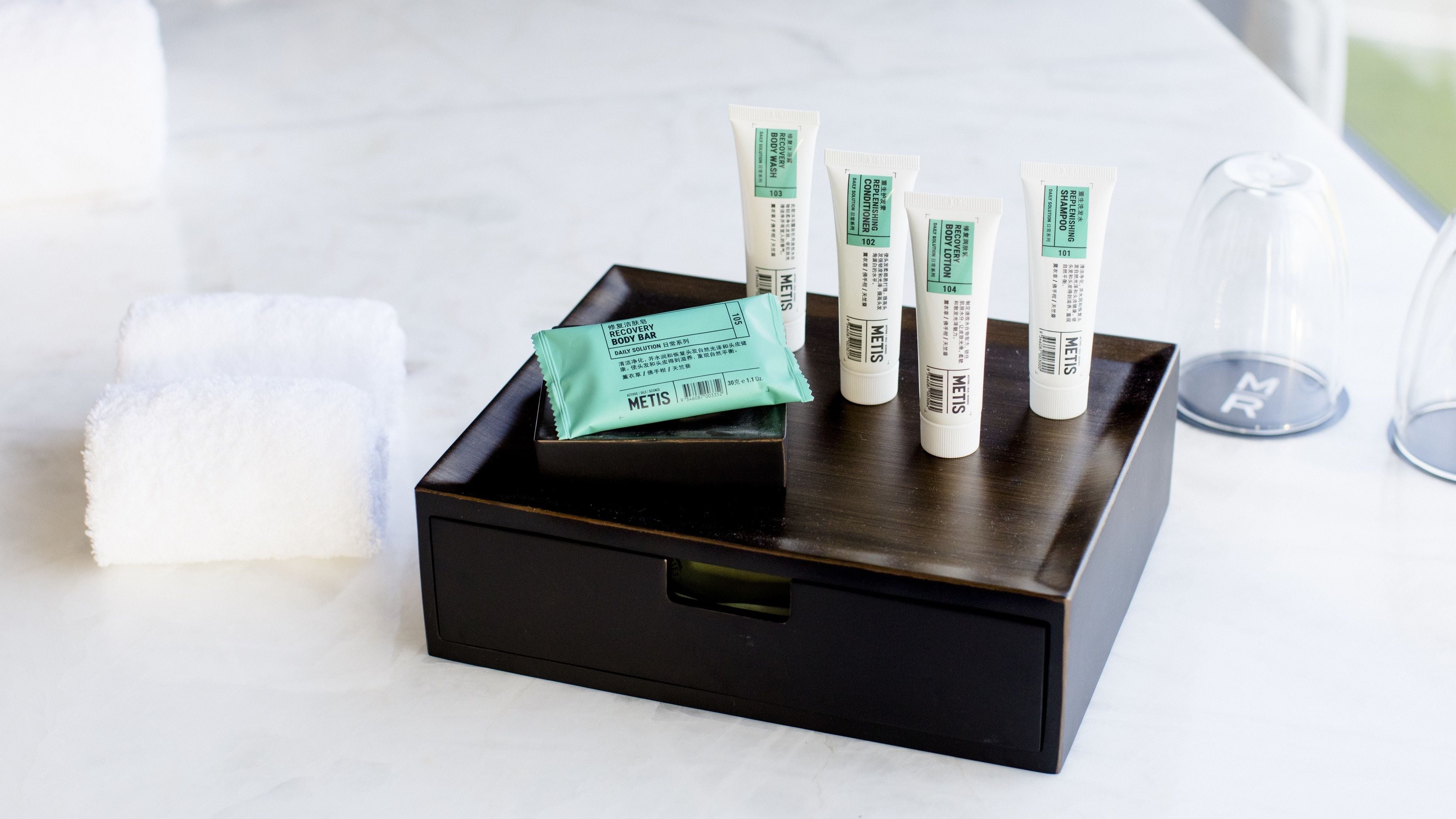 A black tray with toiletries including a bar of soap, several tubes of lotion, and a bar of shampoo on a white marble surface.