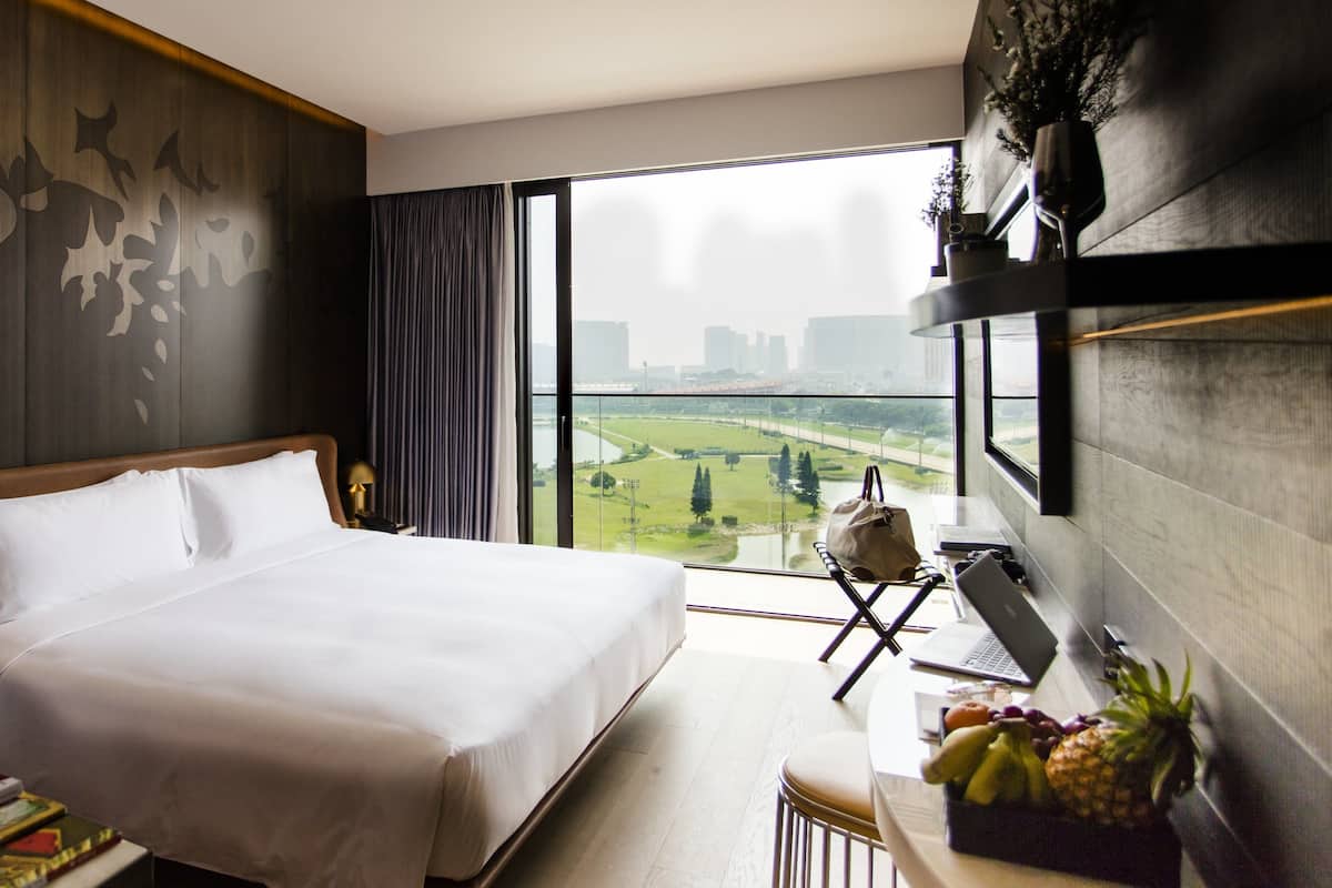A hotel room with a large bed, a desk with a laptop, and a view of a cityscape through a large window.