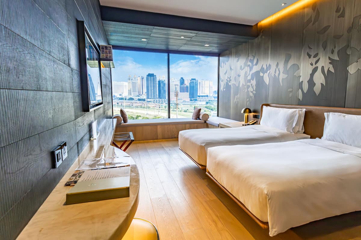 A modern hotel room with two beds, a desk, and a large window offering a city view.
