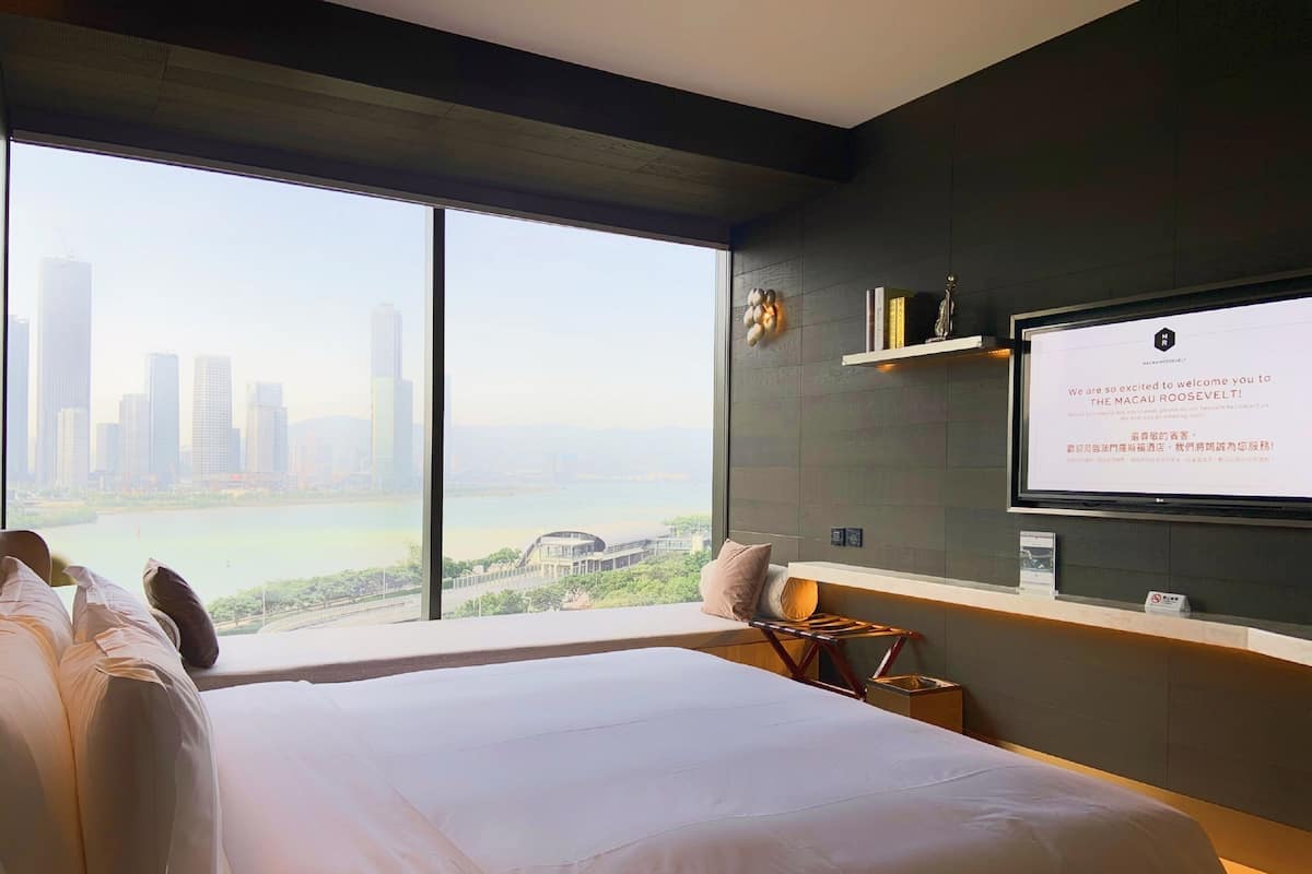 A modern hotel room with a large bed, a TV mounted on the wall, and a view of a city skyline through large windows.