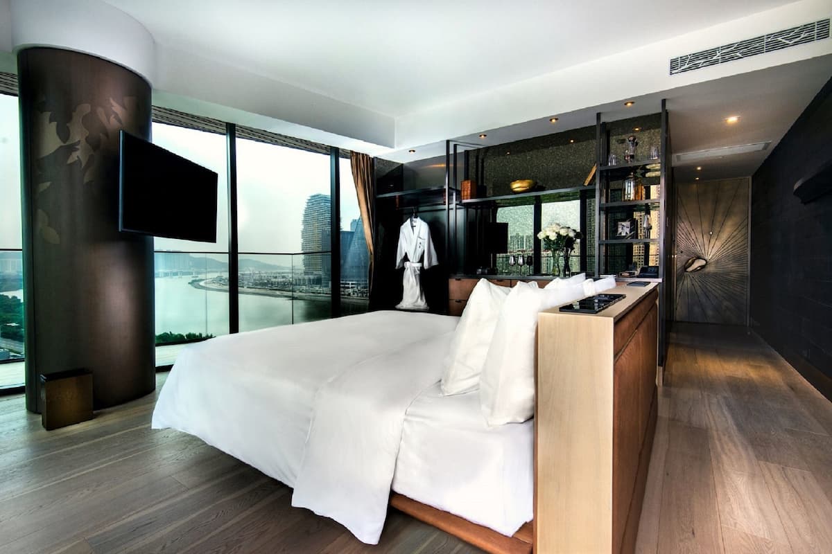 A modern hotel room with a large bed, a TV mounted on a pillar, a view of the cityscape, and a built-in bookshelf.