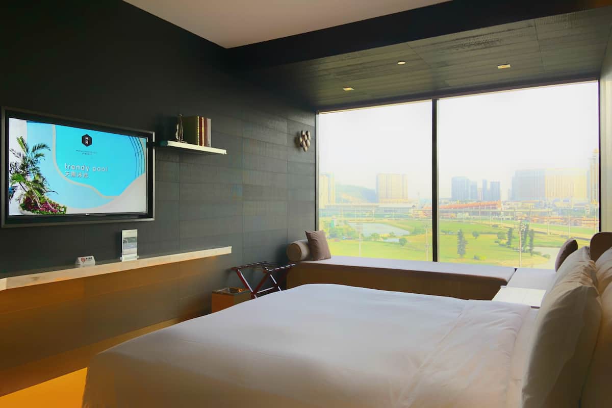 A hotel room with a large bed, a TV on the wall, a window with a city view, and a small table with a lamp.