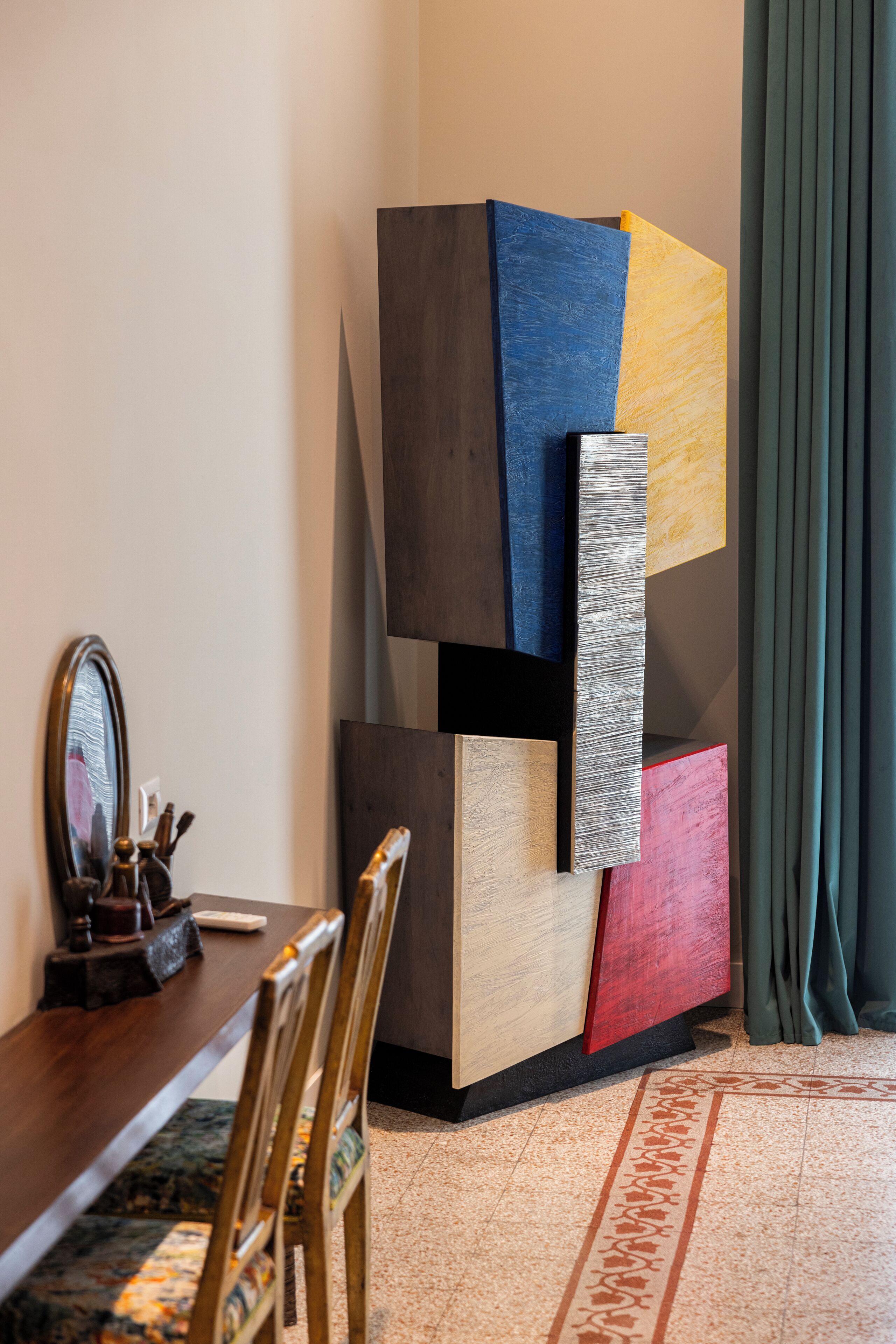 A modern dining area with a wooden table, chairs, and a decorative mirror. A colorful abstract sculpture is mounted on the wall.
