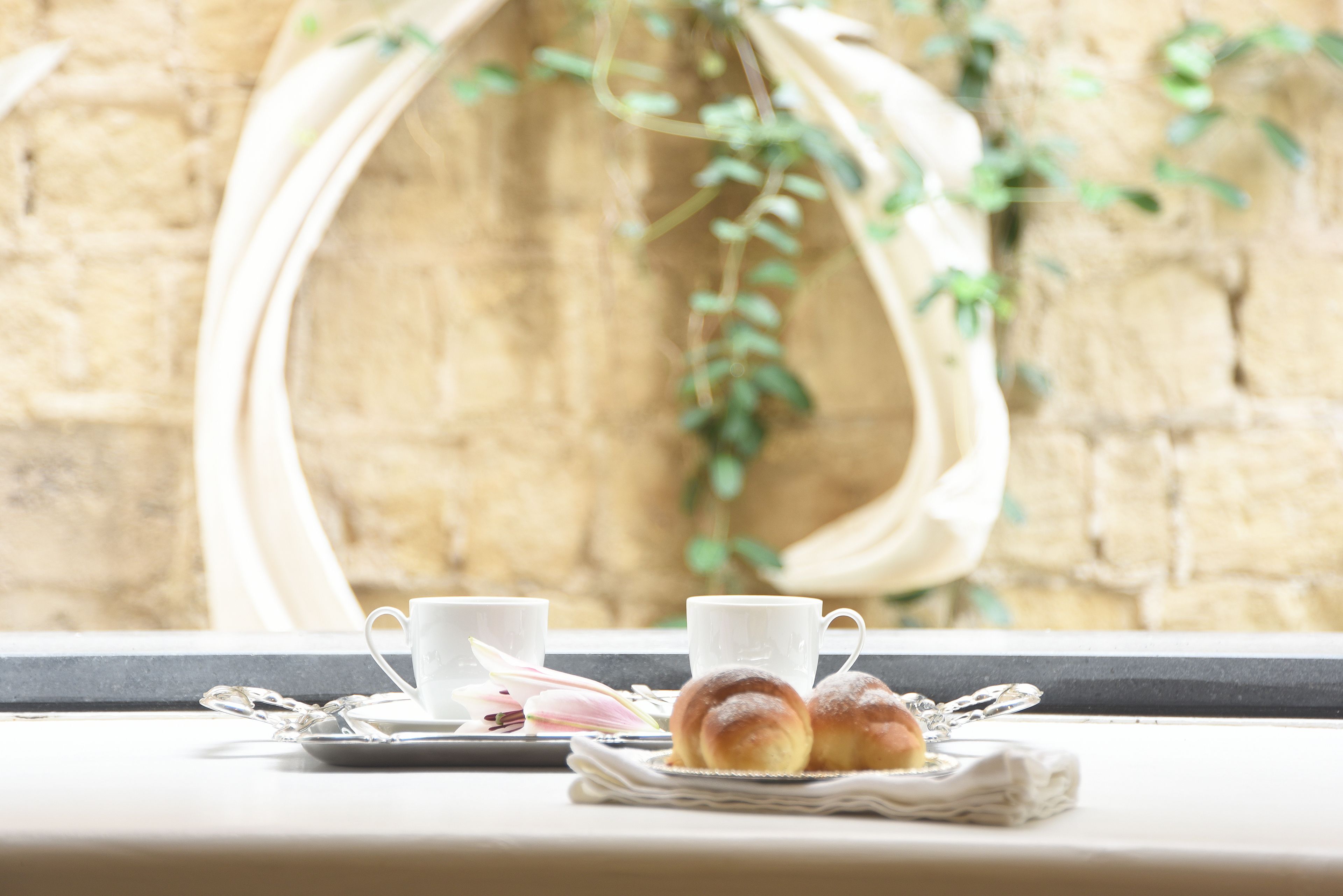 A breakfast setup with two cups, a plate with pastries, and a window with a view of a brick wall and greenery.