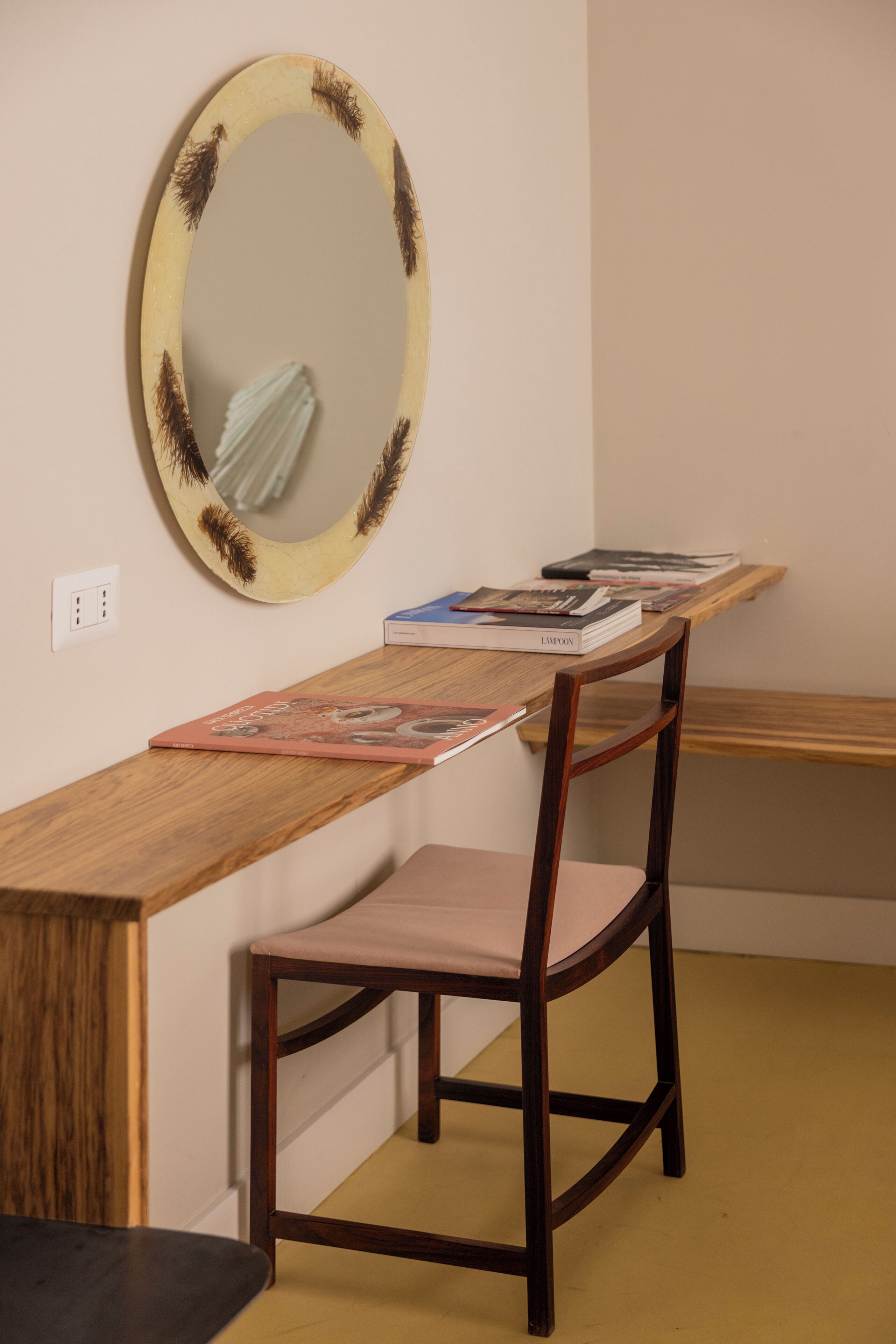 A wooden desk with a chair, a round mirror with a feathered frame, and a shelf with books.