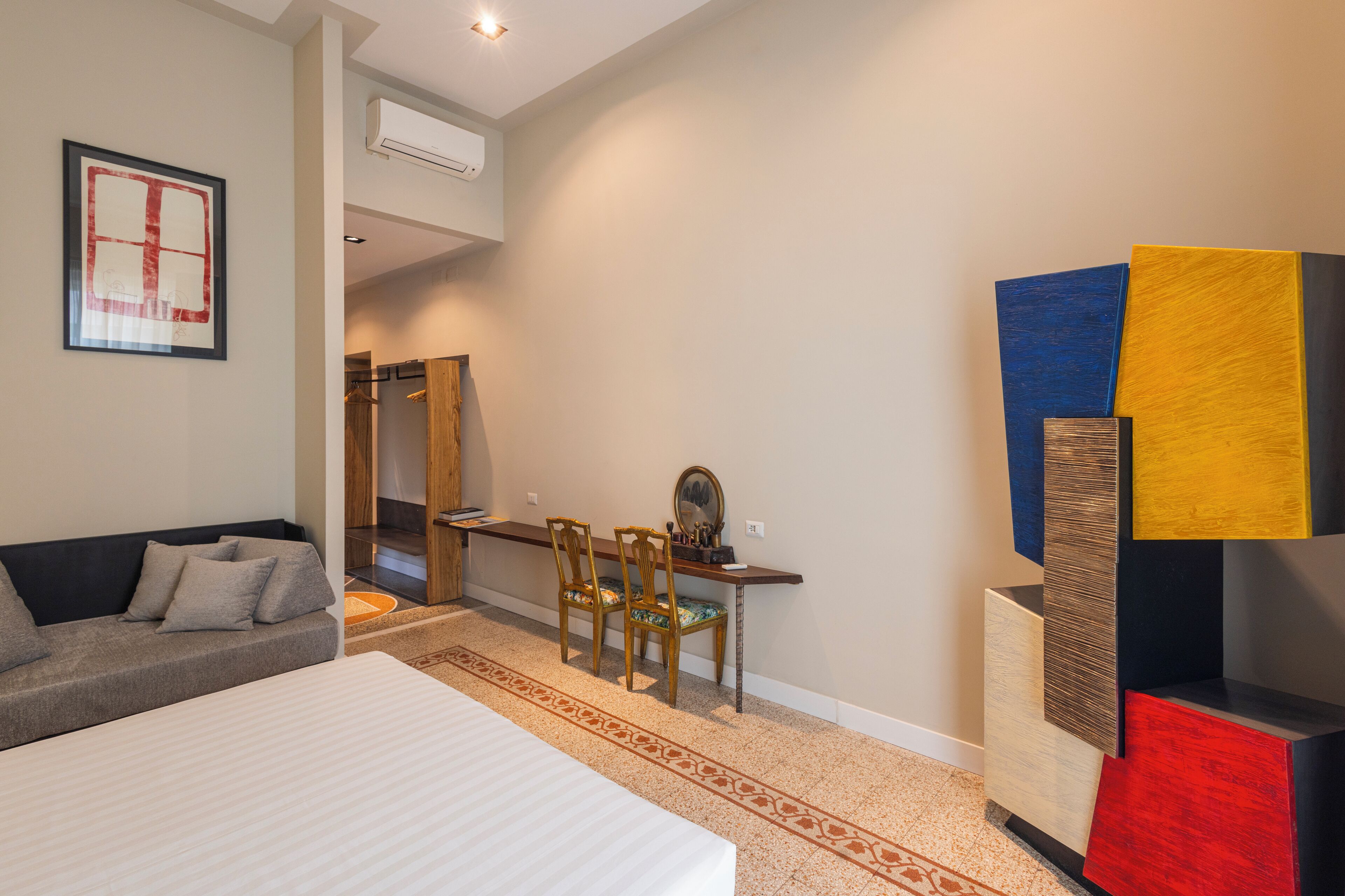 A modern hotel room with a bed, a sofa, a desk with chairs, and a colorful abstract art piece.