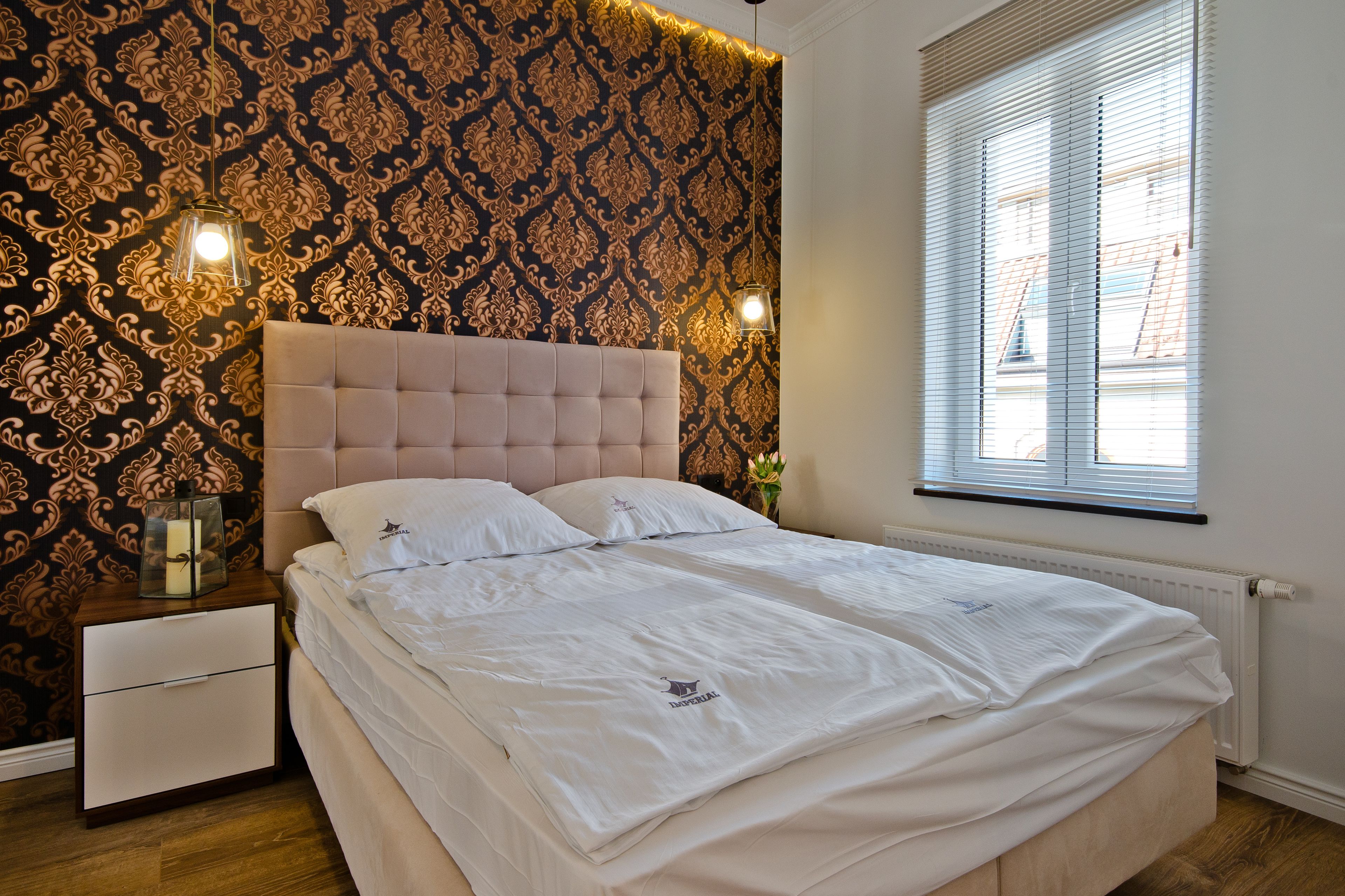 A neatly made bed with a headboard, a bedside table with a lamp, and a patterned wallpaper.