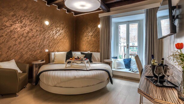 Una camera da letto moderna con un letto rotondo, un comodino con un vassoio, una poltrona e una finestra con tende. C'è anche una TV montata a parete.