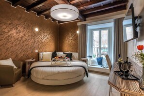 A modern bedroom with a circular bed, a bedside table with a tray, a sofa, a window with curtains, and a TV mounted on the wall. - Spagna Luxury'n Trevi (Rome)