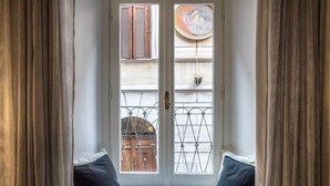 Una stanza con una finestra che si affaccia su un cortile interno, con un tavolino con pasticcini e una tazza, due cuscini scuri, e vista su un edificio con una porta e un cancello in metallo.