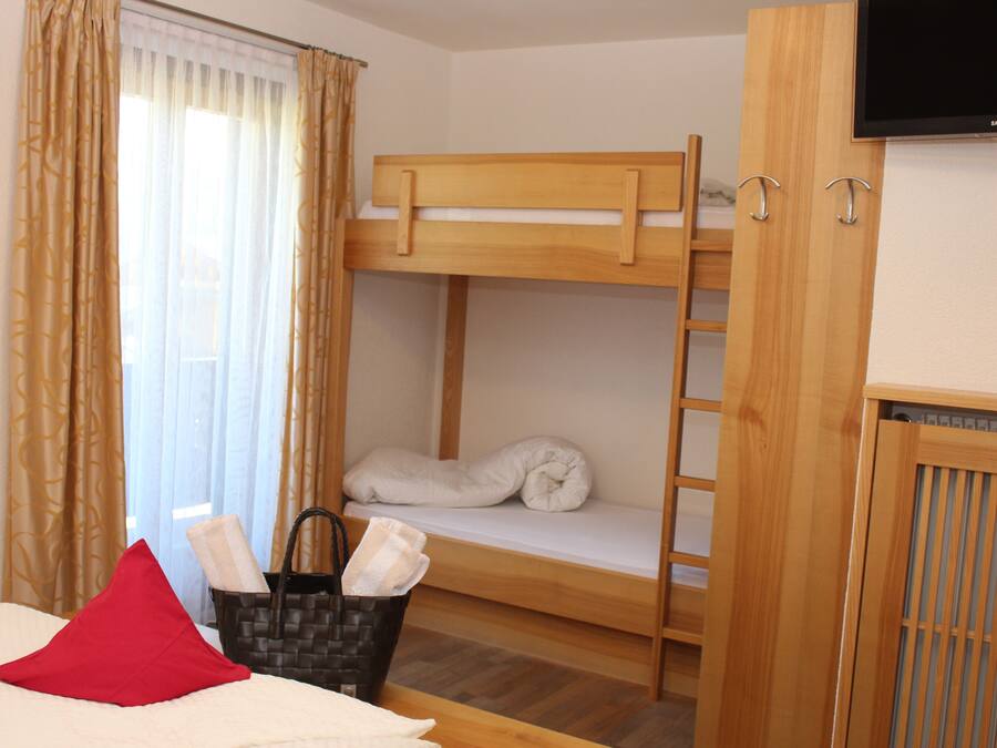 Une chambre avec un lit superposé, une télévision, une fenêtre avec des rideaux, une armoire en bois et une table de chevet avec un oreiller rouge.