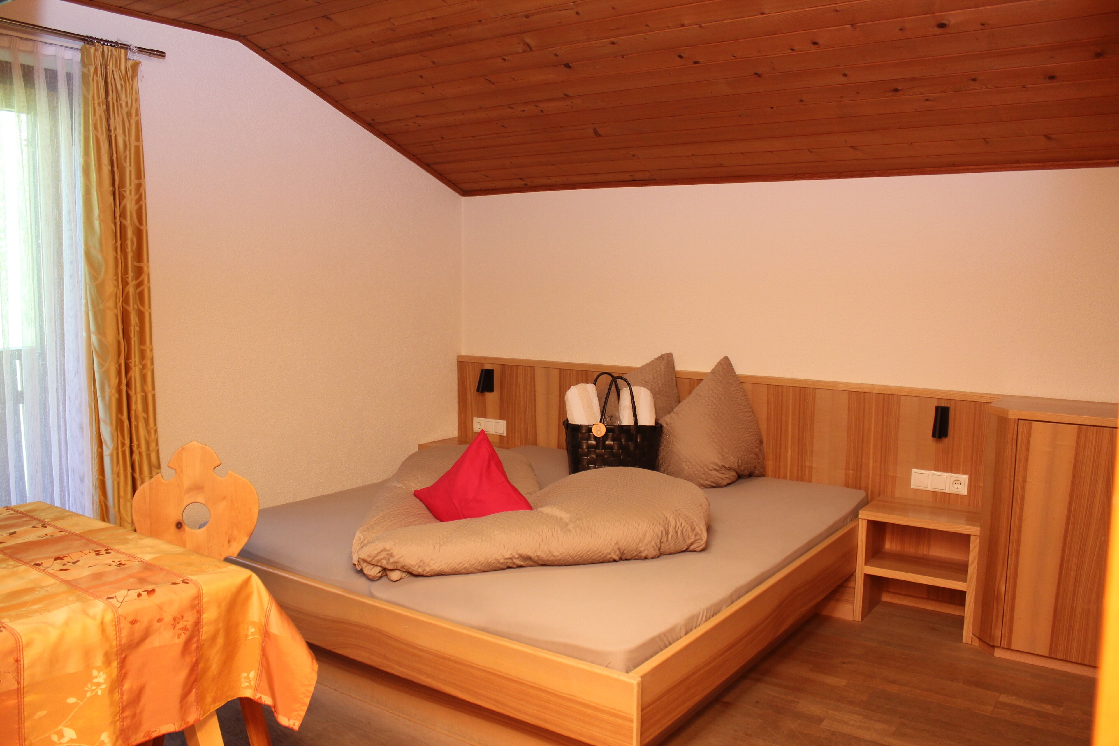 Una cama individual con cabecera de madera, una mesita de noche y un pequeño mueble auxiliar, junto a una ventana con cortinas.