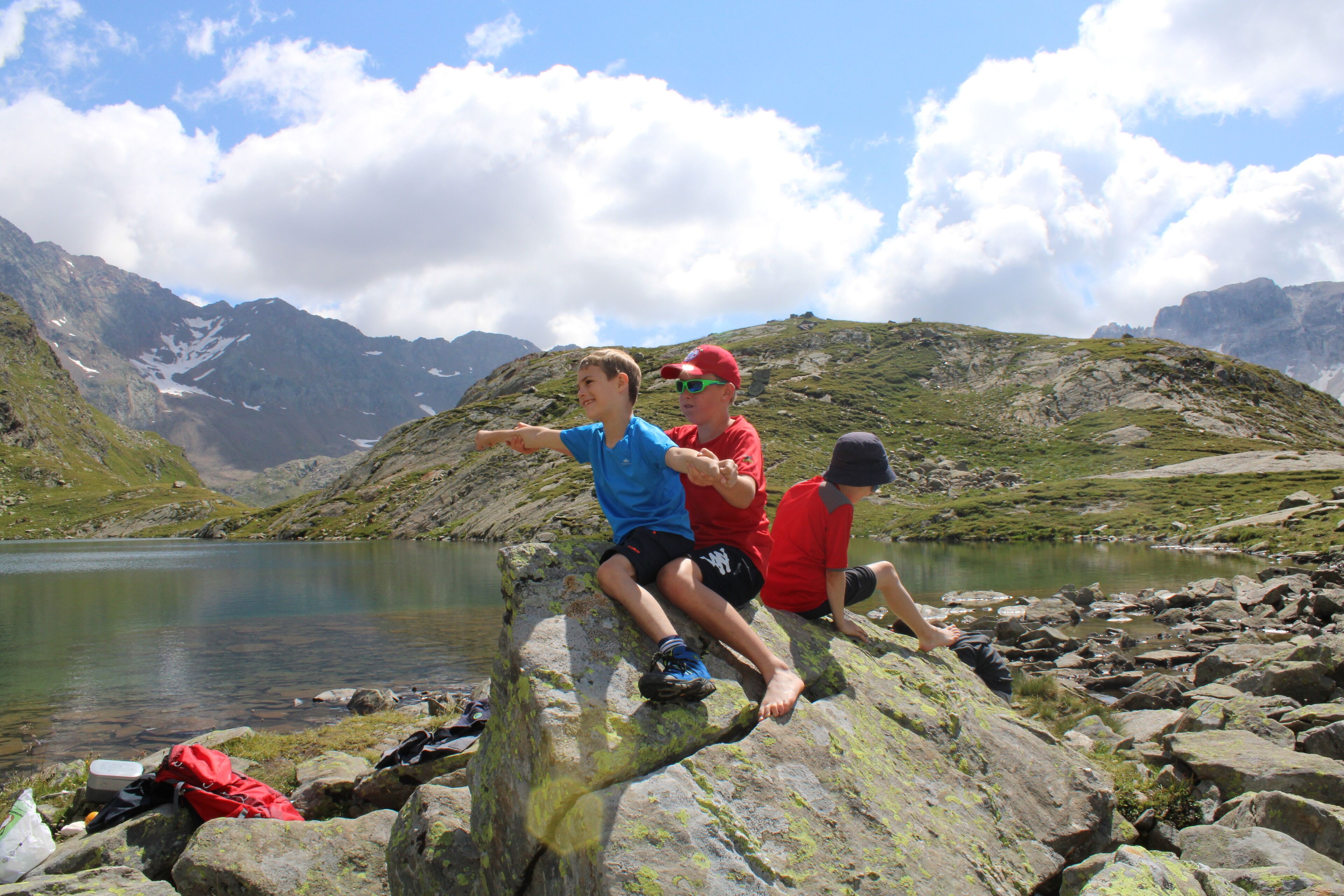 Three people sitting on rocks by a lake with mountains in the background.