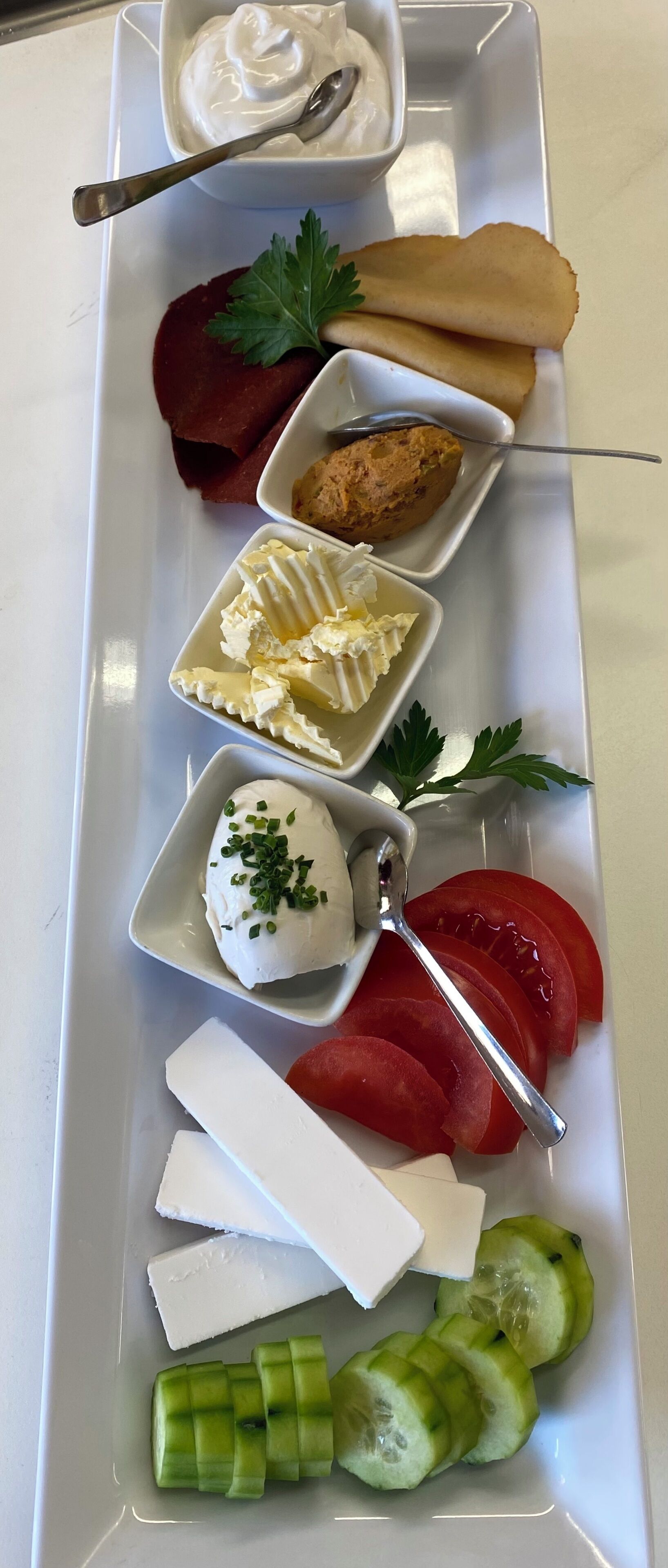 A white rectangular plate with various food items including a dollop of cream, a slice of bread, a piece of meat, a small bowl of sauce, a dollop of cream with chives, a slice of tomato, a piece of cheese, and cucumber slices.