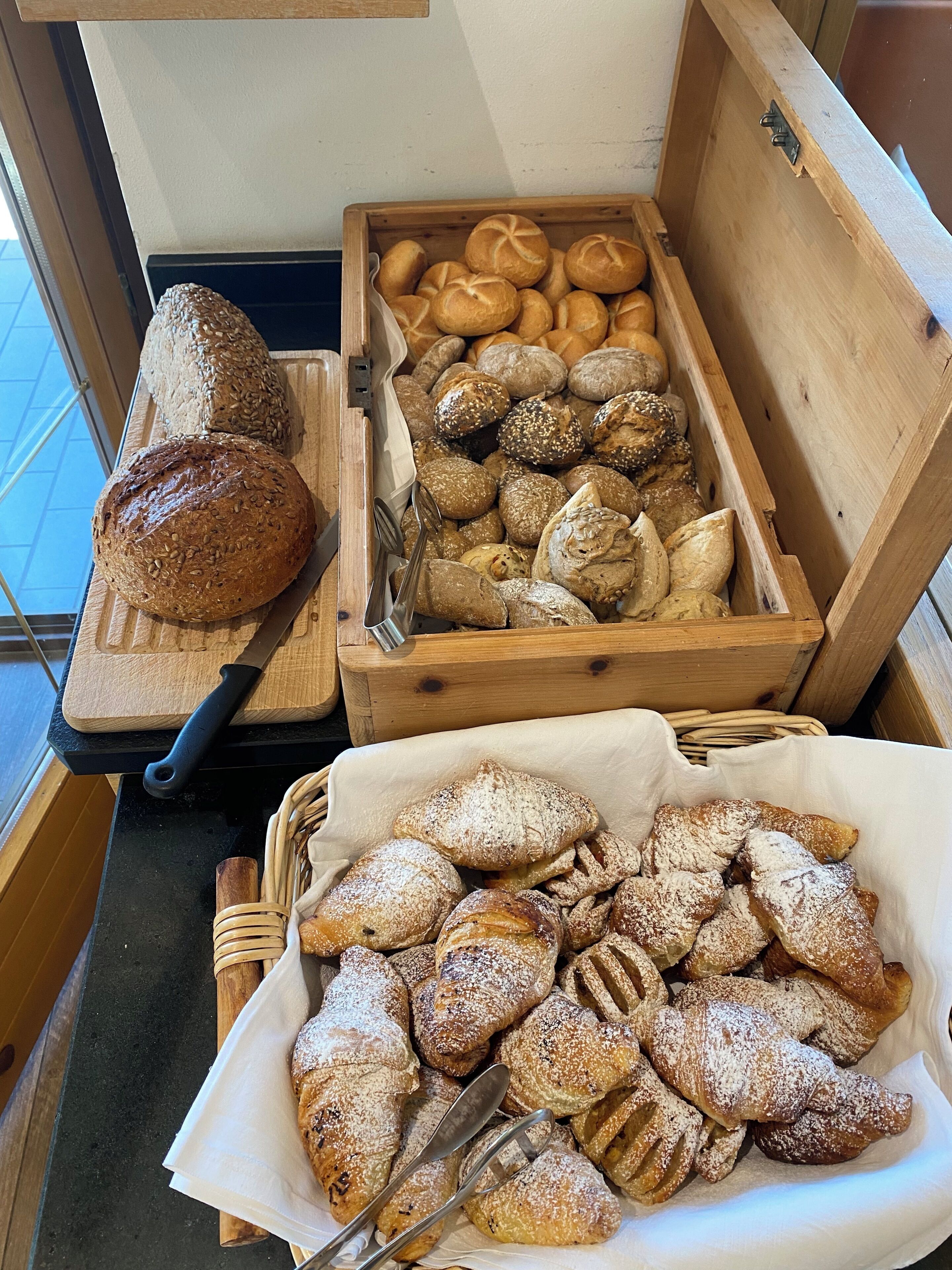 Una caja de madera con panes y pasteles varios, una tabla de cortar con una hogaza de pan, y una cesta de pasteles sobre una encimera.