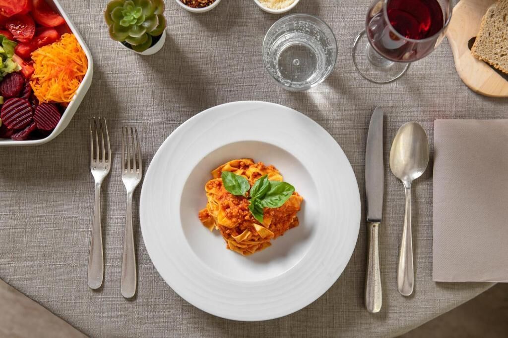 A plate of pasta with a basil garnish, accompanied by a glass of red wine, a bowl of assorted vegetables, and a small succulent plant on a tablecloth.