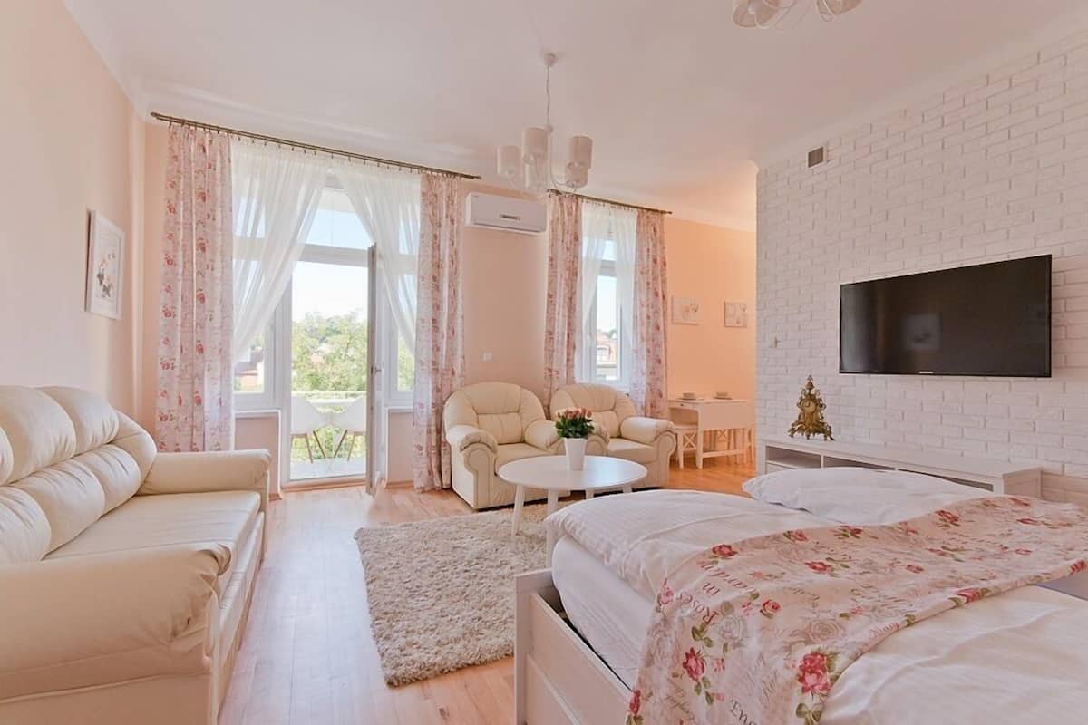 A bedroom with a bed, two chairs, a small table, a TV, and a balcony with curtains.