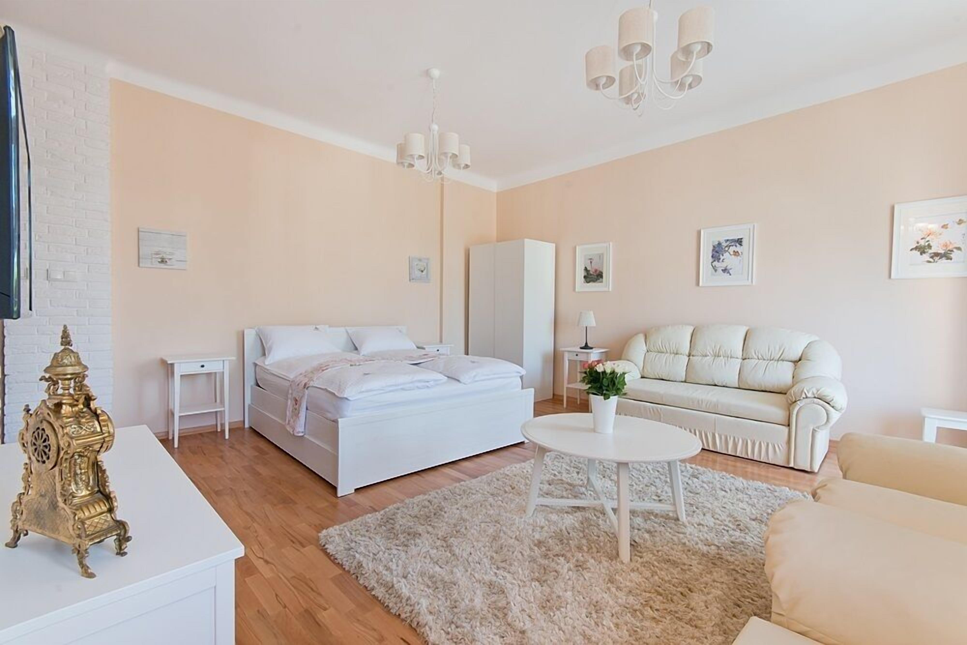 Un dormitorio moderno con cama, mesitas de noche, un tocador blanco, una alfombra mullida, un sofá blanco y una araña.