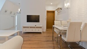 A modern, well-lit room with a wooden floor, white furniture, a flat-screen TV mounted on the wall, and a dining area with a table and chairs.