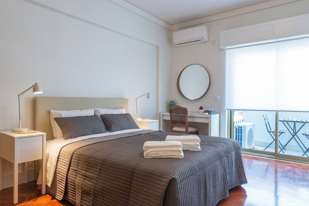 A bedroom with a bed, a desk, a chair, a round mirror, and a balcony with a table and chairs.