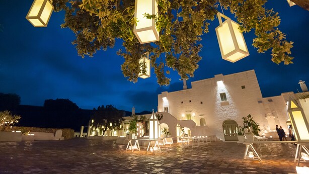 Un patio con faroles colgantes, un edificio blanco con arcos y suelo de piedra.
