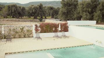 A pool area with lounge chairs, a stone wall, and a view of olive trees and hills.