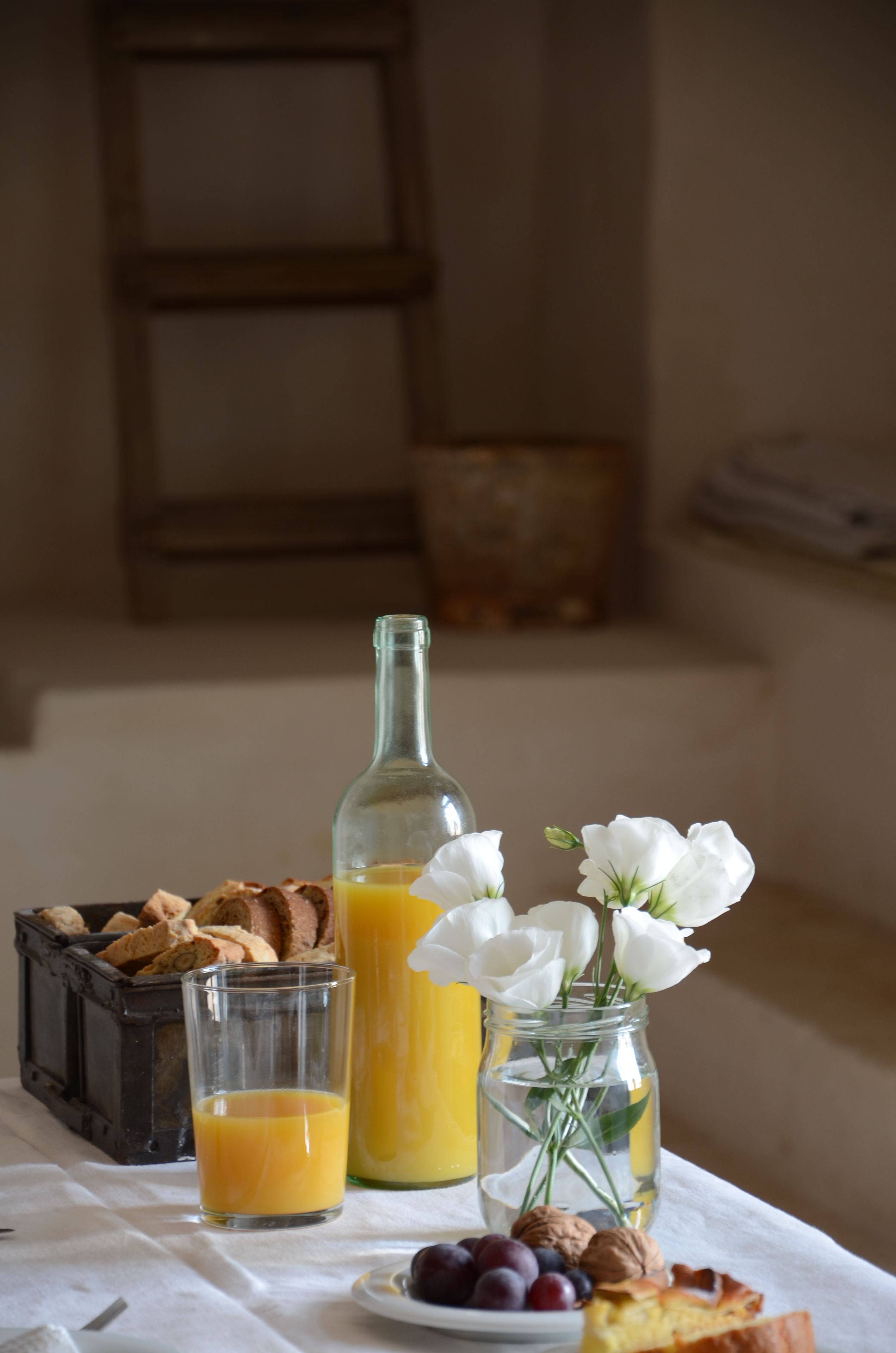 A breakfast setting with a glass of orange juice, a bottle of orange juice, a jar of flowers, a plate of grapes, and a slice of cake on a table.