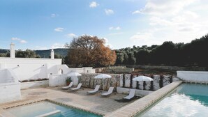 Zona de piscina con sillones de descanso y sombrillas, rodeada de paredes blancas y un muro de piedra con vegetación.