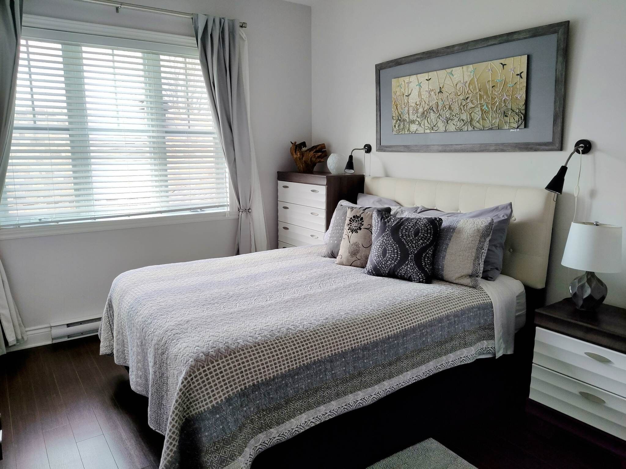 A bedroom with a bed, a dresser, a nightstand, a lamp, a window with blinds, and a framed artwork on the wall.