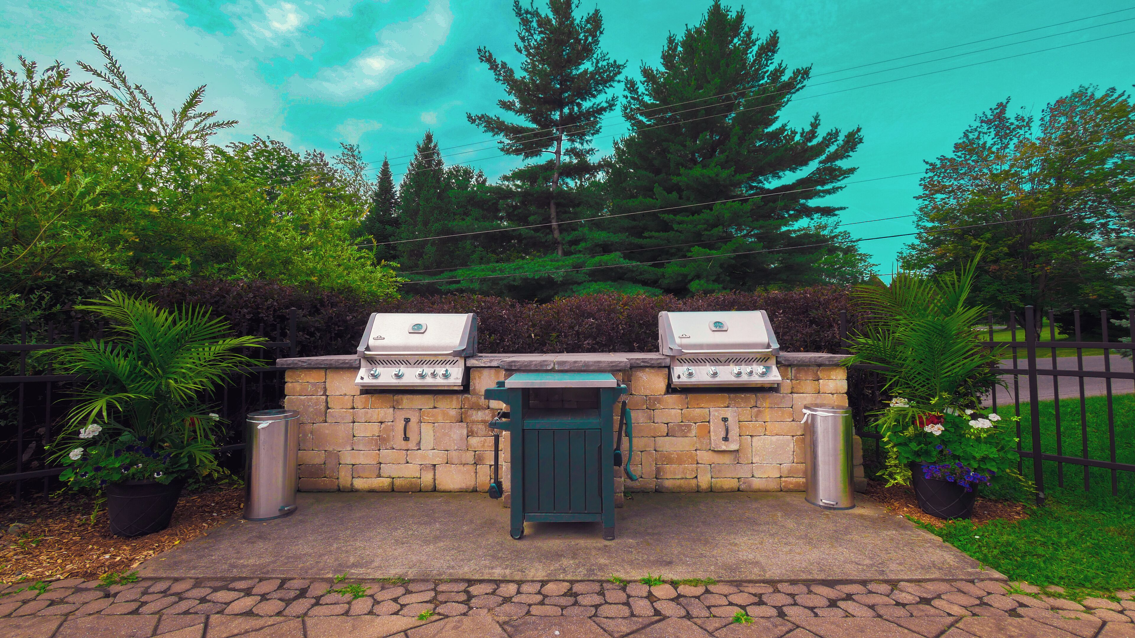 An outdoor barbecue area with two grills, a green cabinet, and a trash can, surrounded by greenery and a stone wall.