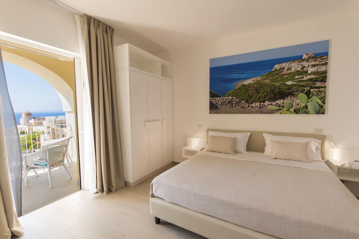 Ein Schlafzimmer mit Bett, Nachttisch, einem Gemälde einer Küstenlandschaft und einem Balkon mit Blick auf Stadt und Meer.