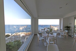 Une chambre située dans un immeuble de grande hauteur, offrant une vue panoramique sur la mer, meublée de chaises et de tables blanches, et agrémentée d’une plante en pot.