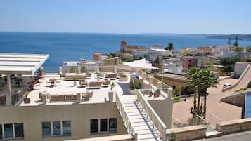Una terraza en la azotea con mobiliario exterior, vista al mar y una combinación de edificios tradicionales y modernos al fondo.
