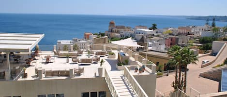 A rooftop terrace with outdoor seating, a view of the sea, and a mix of traditional and modern buildings in the background.