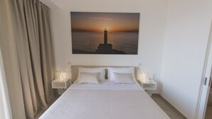 A hotel room with a large bed, two bedside tables with lamps, a painting of a lighthouse on the wall, and a view of the sea. - Est Hotel (Santa Cesarea Terme)