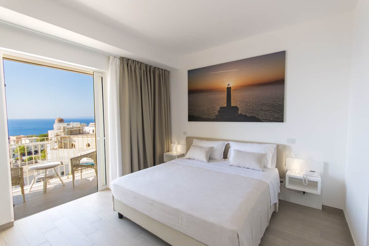 Ein Hotelzimmer mit einem großen Bett, einem Nachttisch, einem Fenster mit Meerblick und einem Gemälde eines Sonnenuntergangs.