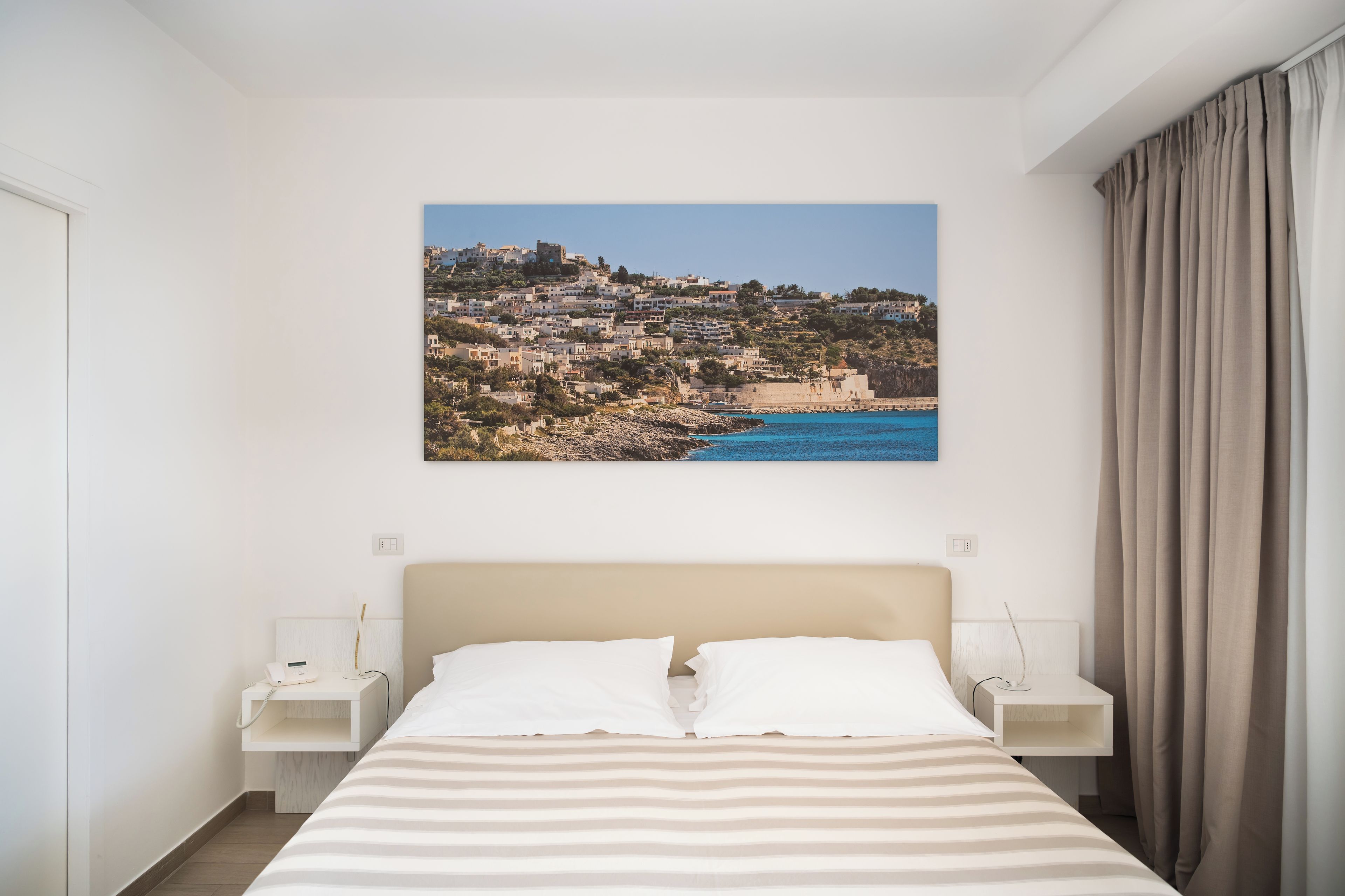 Una habitación de hotel con una cama, dos mesitas de noche, un cabecero, un cuadro de un pueblo costero y una cortina.