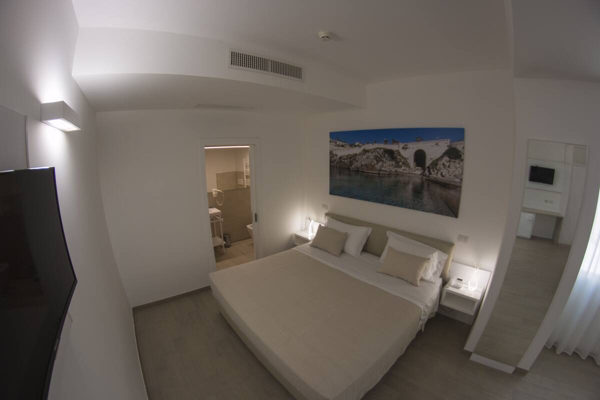 Ein Schlafzimmer mit einem großen Bett, einem Gemälde einer Küstenlandschaft, einem Fernseher und einem durch eine offene Tür einsehbaren Badezimmer.