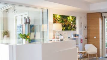 Un espace d’accueil avec un comptoir en verre, une chaise blanche, une lampe, un arrangement floral fixé au mur et un miroir qui reflète l’intérieur.