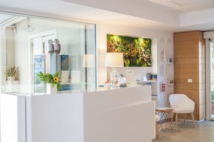 A reception area with a glass counter, a white chair, a lamp, a wall-mounted flower display, and a mirror reflecting the interior.