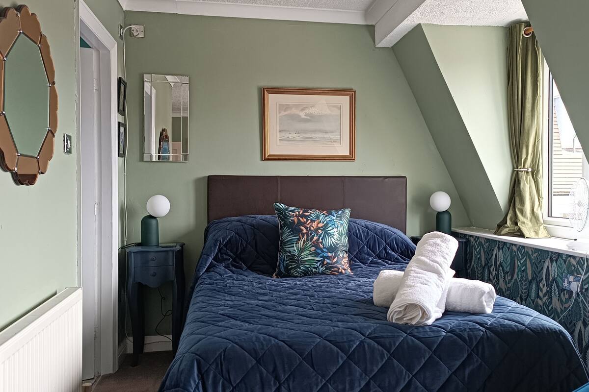 Quarto com cama, mesa de cabeceira, espelho, quadro emoldurado e janela com cortinas.